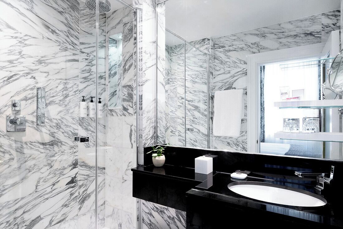 A modern bathroom with a marble wall, a glass shower enclosure, a black countertop, and a white sink.