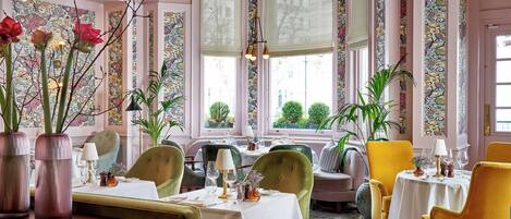 A dining area with tables set for two, featuring green and yellow upholstered chairs, a chandelier, and large windows with patterned curtains.