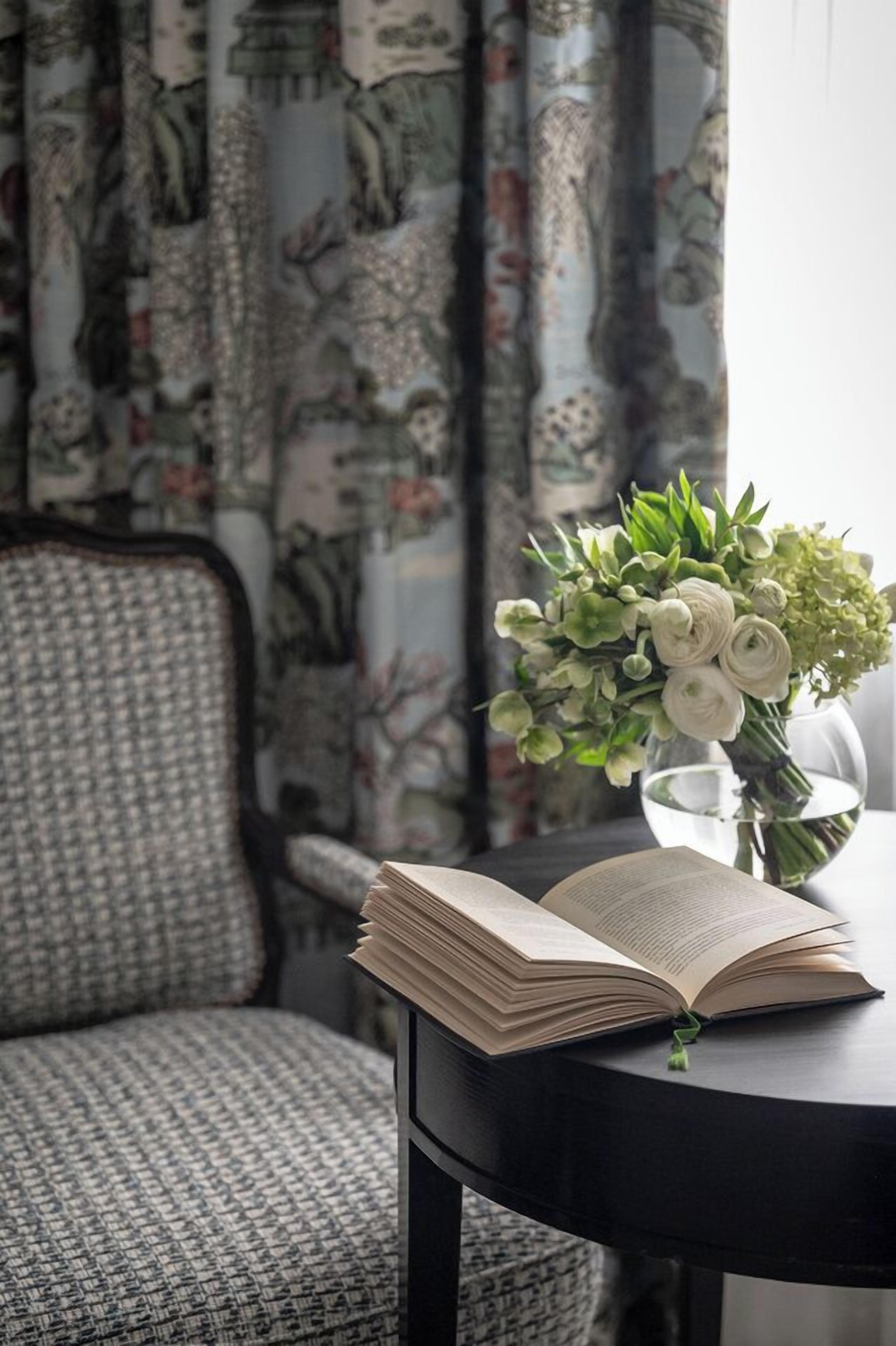 Ein offenes Buch auf einem schwarzen Tisch mit einem grünen Lesezeichen, eine Vase mit Blumen und ein Stuhl mit gemustertem Stoff.