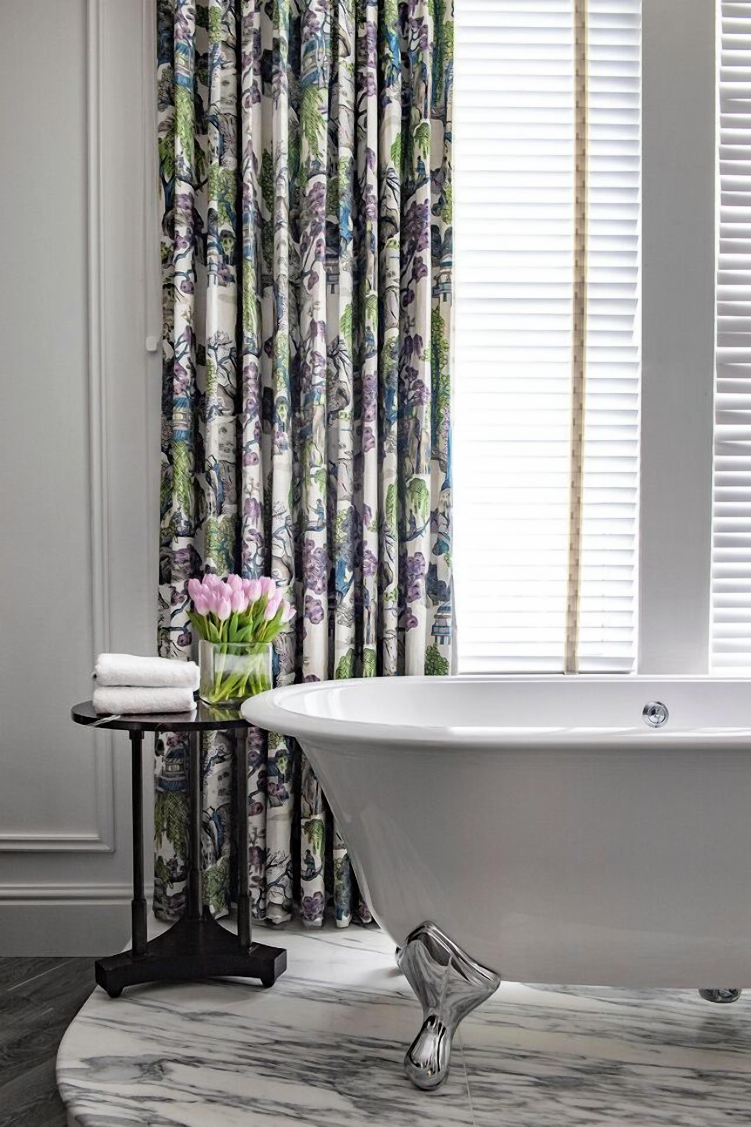 A bathroom with a freestanding bathtub, a small table with a vase of flowers, and a window with blinds.