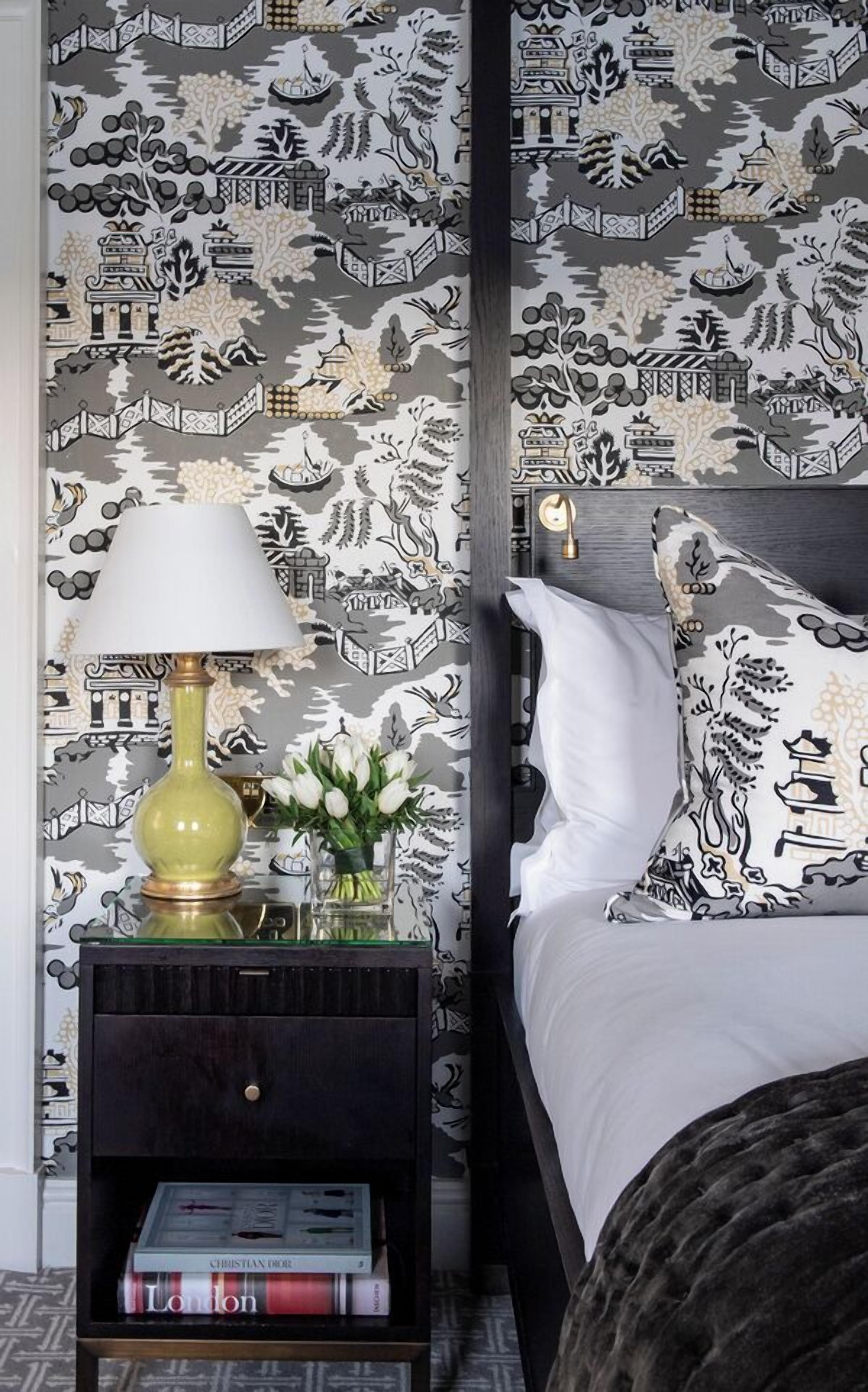 A bedroom with a patterned wallpaper, a bedside table with a lamp, a vase with flowers, and a bed with pillows.