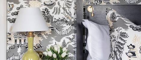 A bedroom with a patterned wallpaper, a bedside table with a lamp, a vase with flowers, and a bed with pillows.