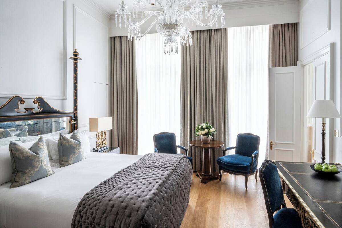 A hotel room with a large bed, a chandelier, two armchairs, a round table with a vase of flowers, and a view of the city through large windows.
