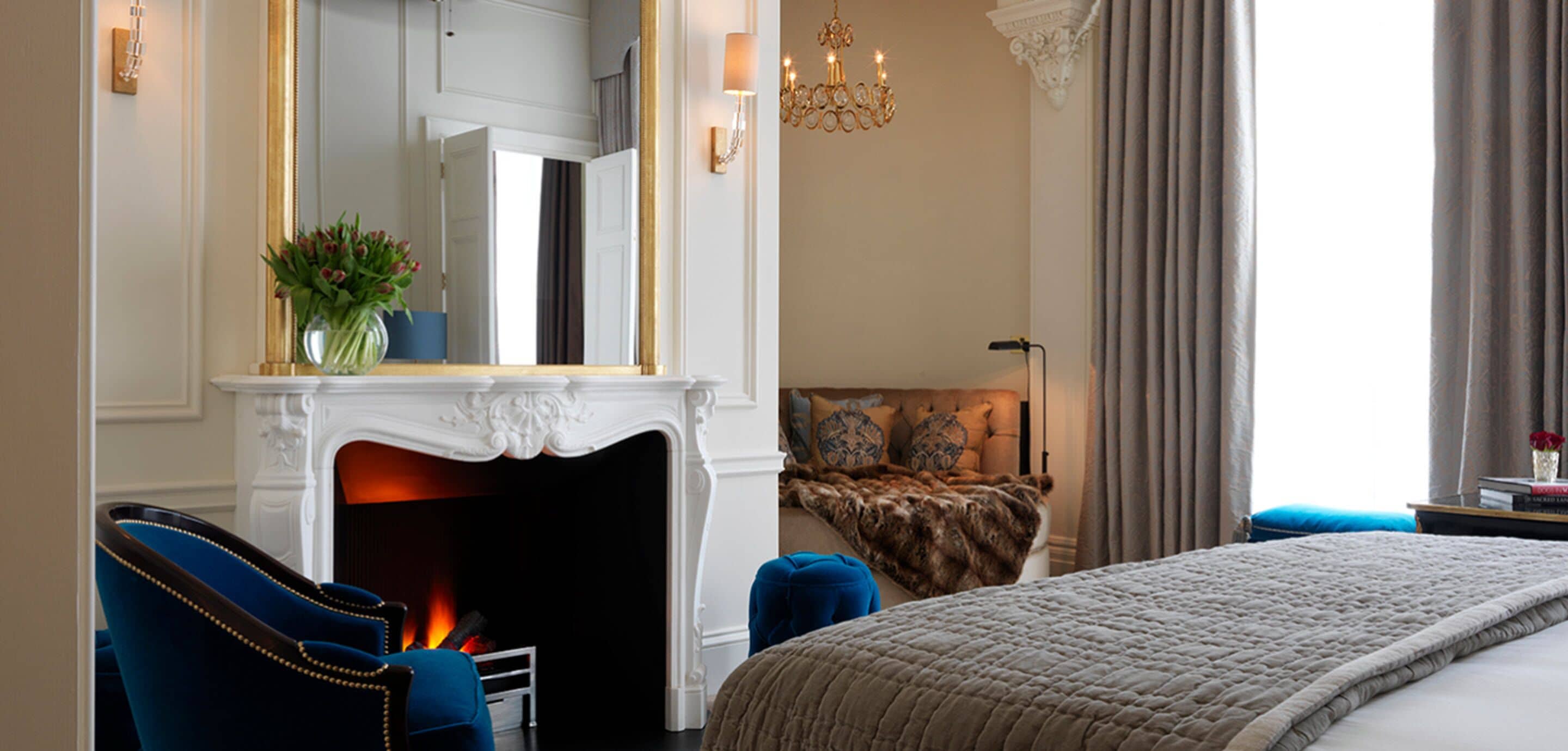 A bedroom with a fireplace, a bed, a blue armchair, and a chandelier.
