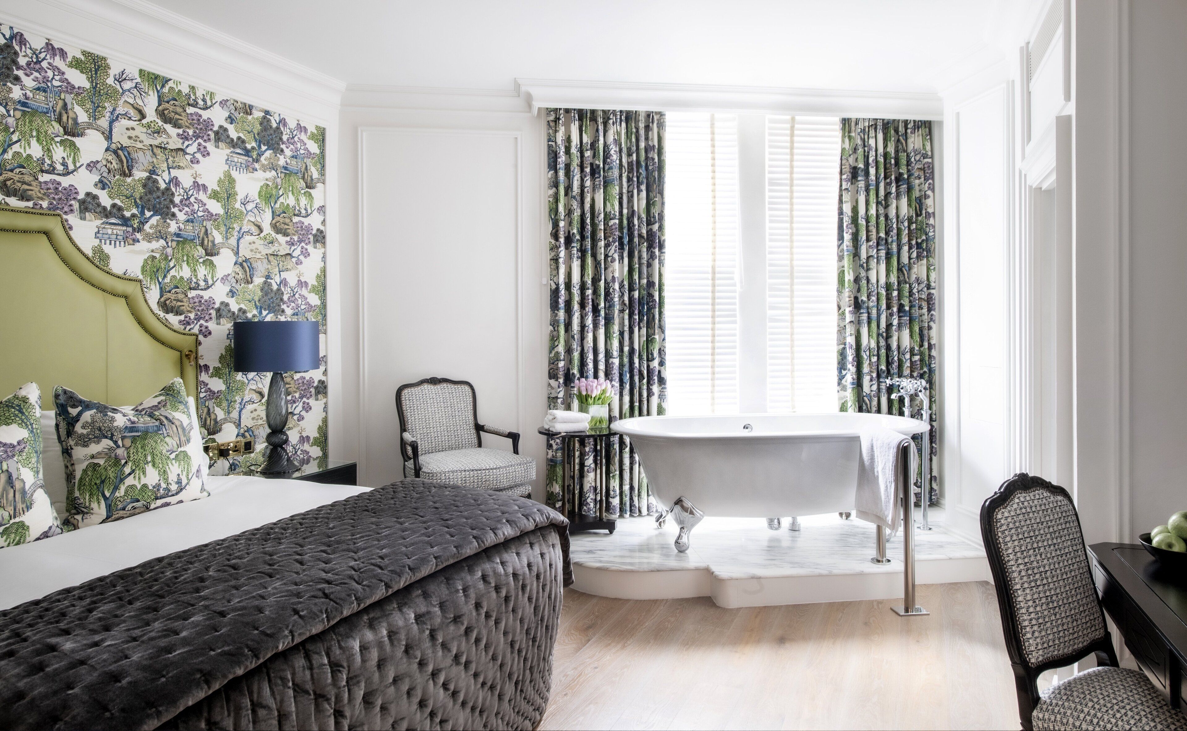 Une chambre à coucher avec un grand lit, une chaise, une petite table, une baignoire et des rideaux ornés d’un motif naturel.