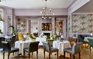 A dining area with multiple round tables, chairs, and a central fireplace. - The Kensington Hotel (London)