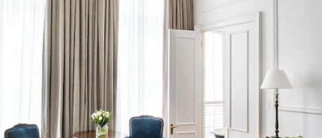 Habitación de hotel con una cama grande, un candelabro, un escritorio con lámpara y vistas al exterior a través de amplias ventanas.