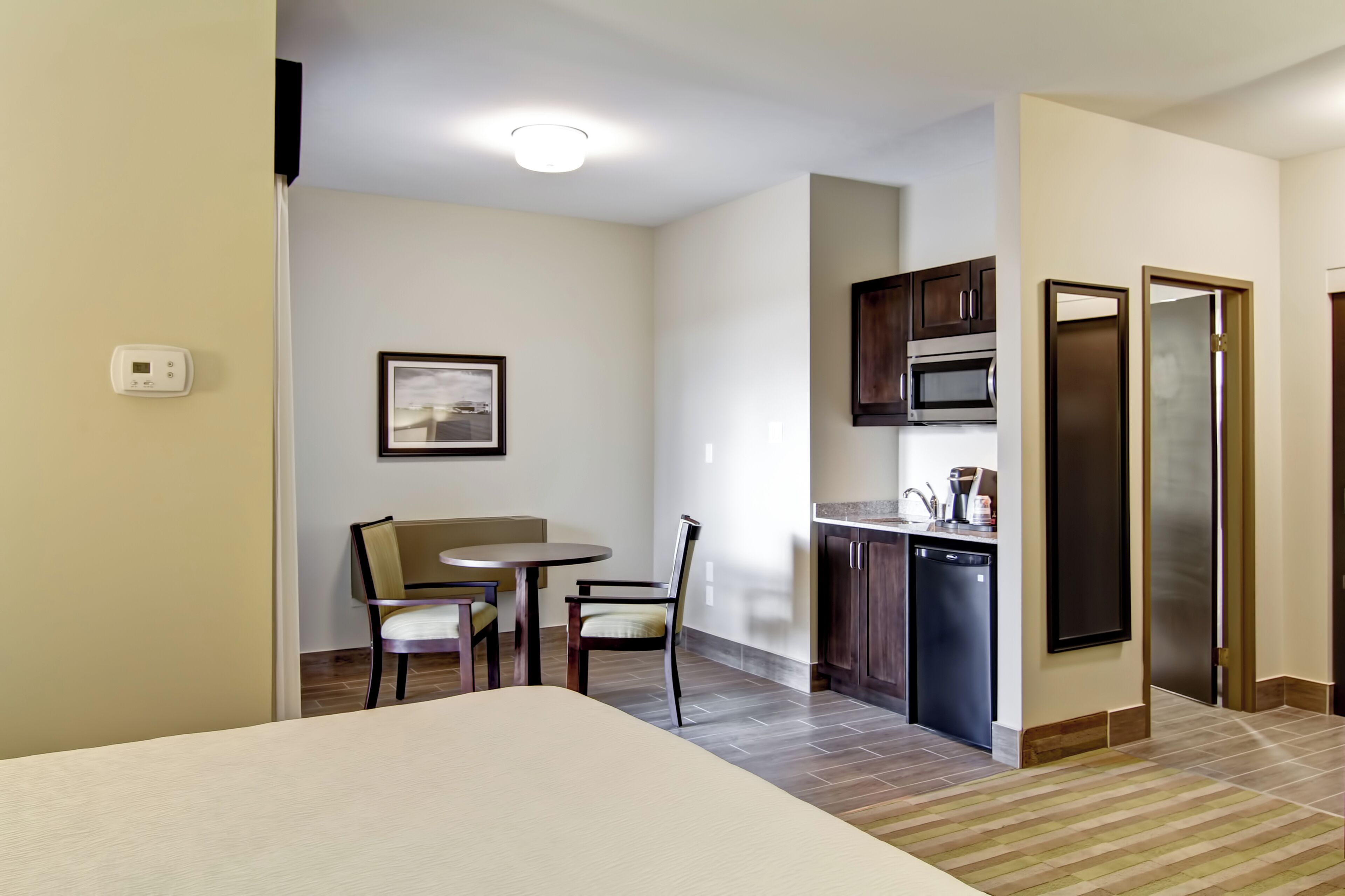 A compact hotel room with a bed, a small dining table with chairs, a kitchenette with a microwave and refrigerator, and a framed picture on the wall.