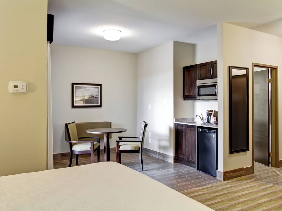 A compact hotel room with a bed, a small dining table with chairs, a kitchenette with a microwave and refrigerator, and a framed picture on the wall.