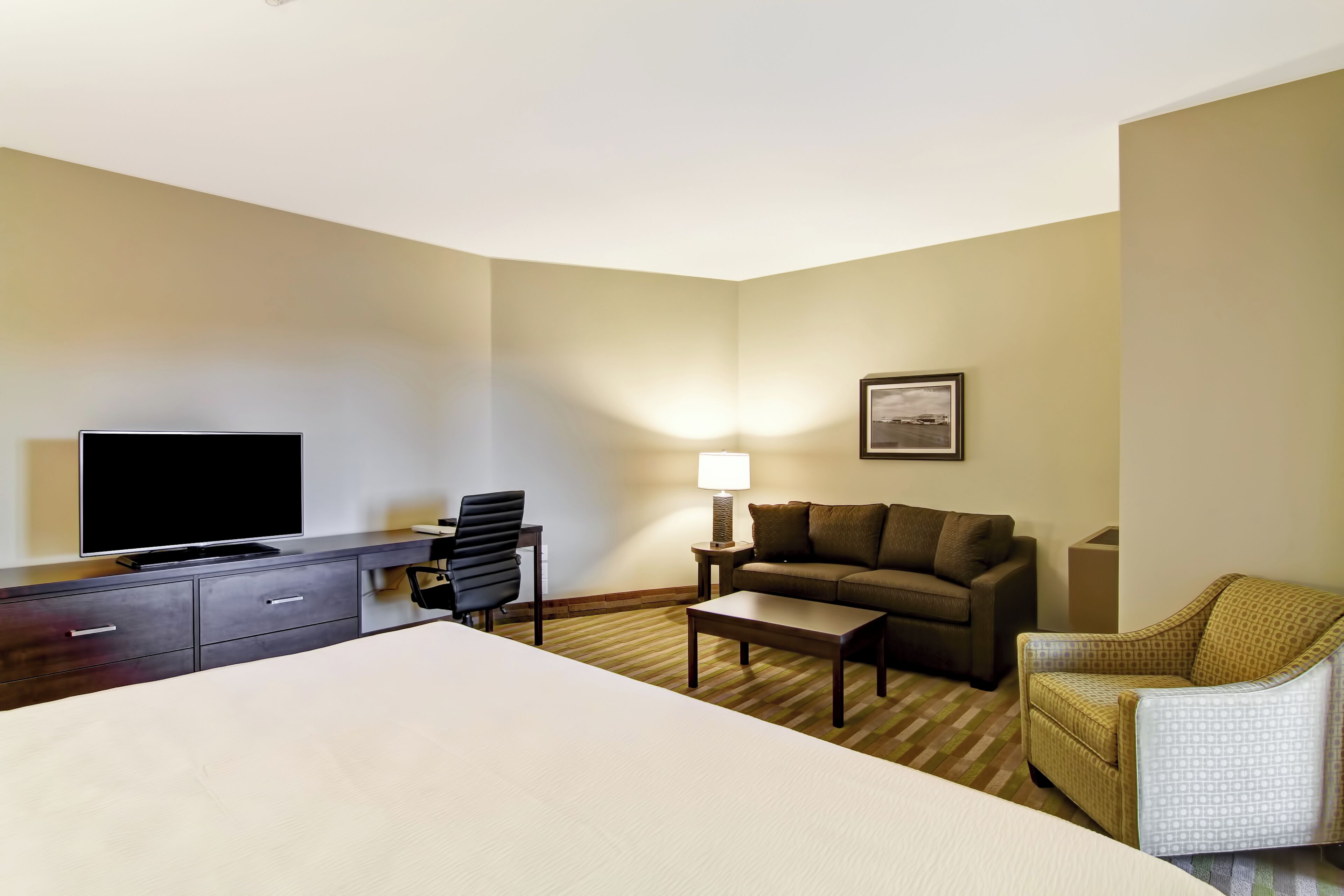 A hotel room with a bed, a desk with a TV, a sofa, a chair, and a lamp.