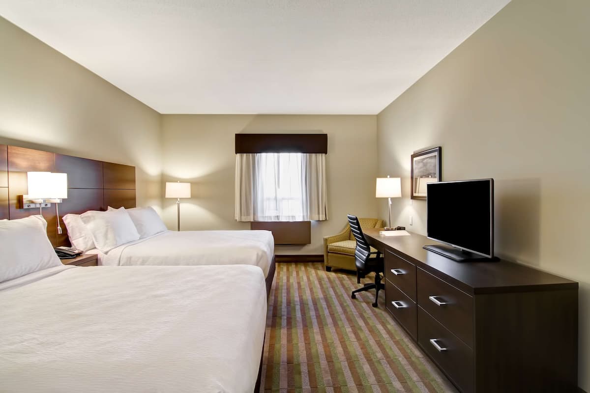 Ein Hotelzimmer mit zwei Betten, einem Schreibtisch mit Fernseher, einem Sessel und einem Fenster mit Vorhängen.