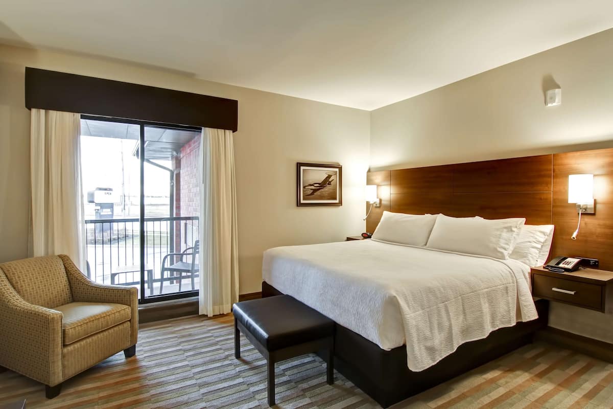 Ein Hotelzimmer mit einem großen Bett, einem Sessel, einer Bank, einem Nachttisch und Blick ins Freie durch eine Schiebetür.