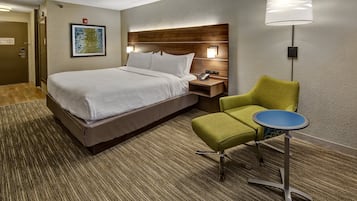 A hotel room with a bed, a green armchair, a small table, and a wall-mounted lamp.