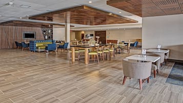 Un vestíbulo de hotel espacioso con varias zonas de estar, incluyendo una mesa de comedor central rodeada de sillas, una zona de sofás y mesas individuales.