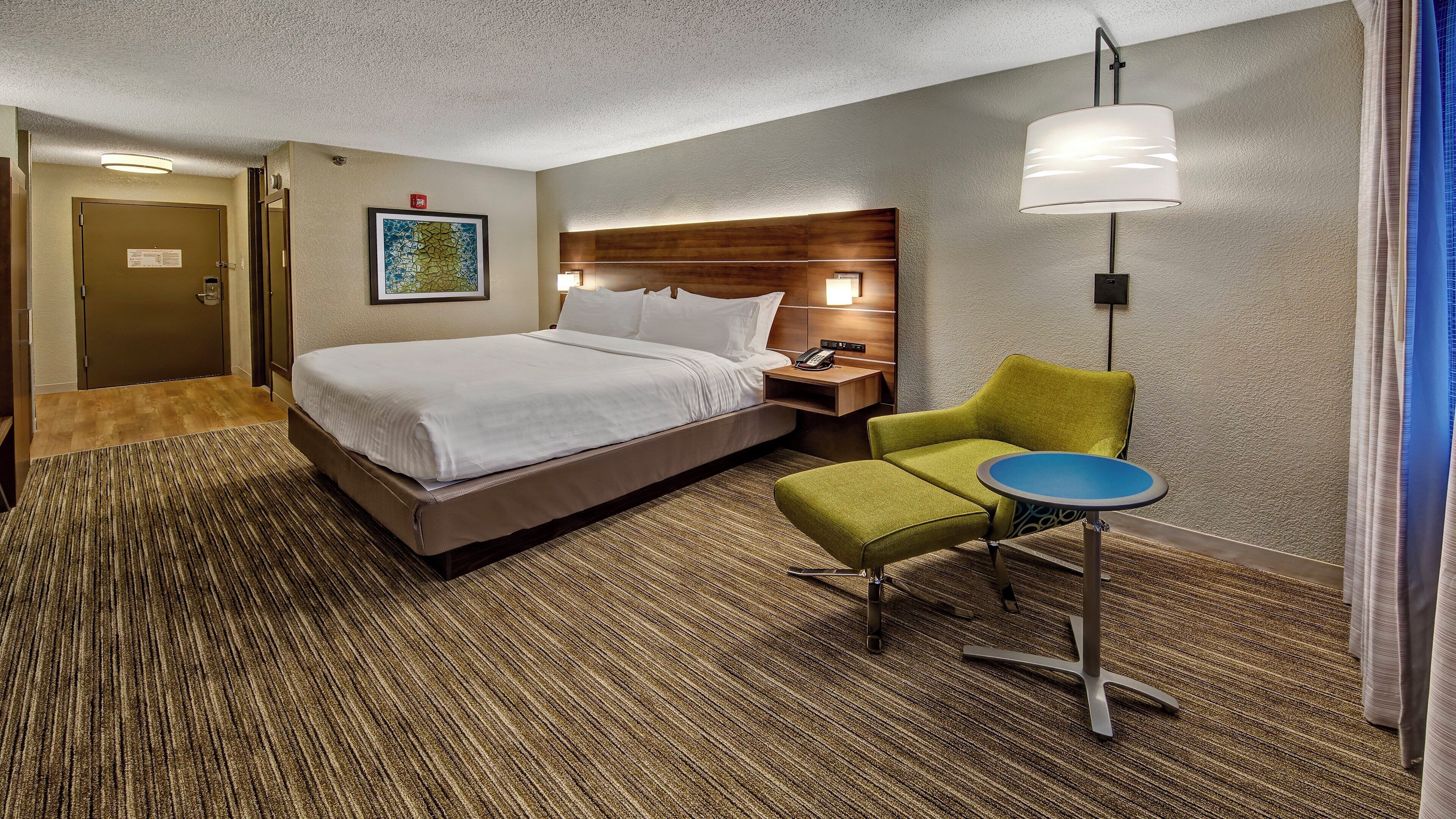 Ein Hotelzimmer mit Bett, einem grünen Sessel, einem kleinen Tisch, einem Nachttisch, einer Wandlampe und einem gerahmten Bild.