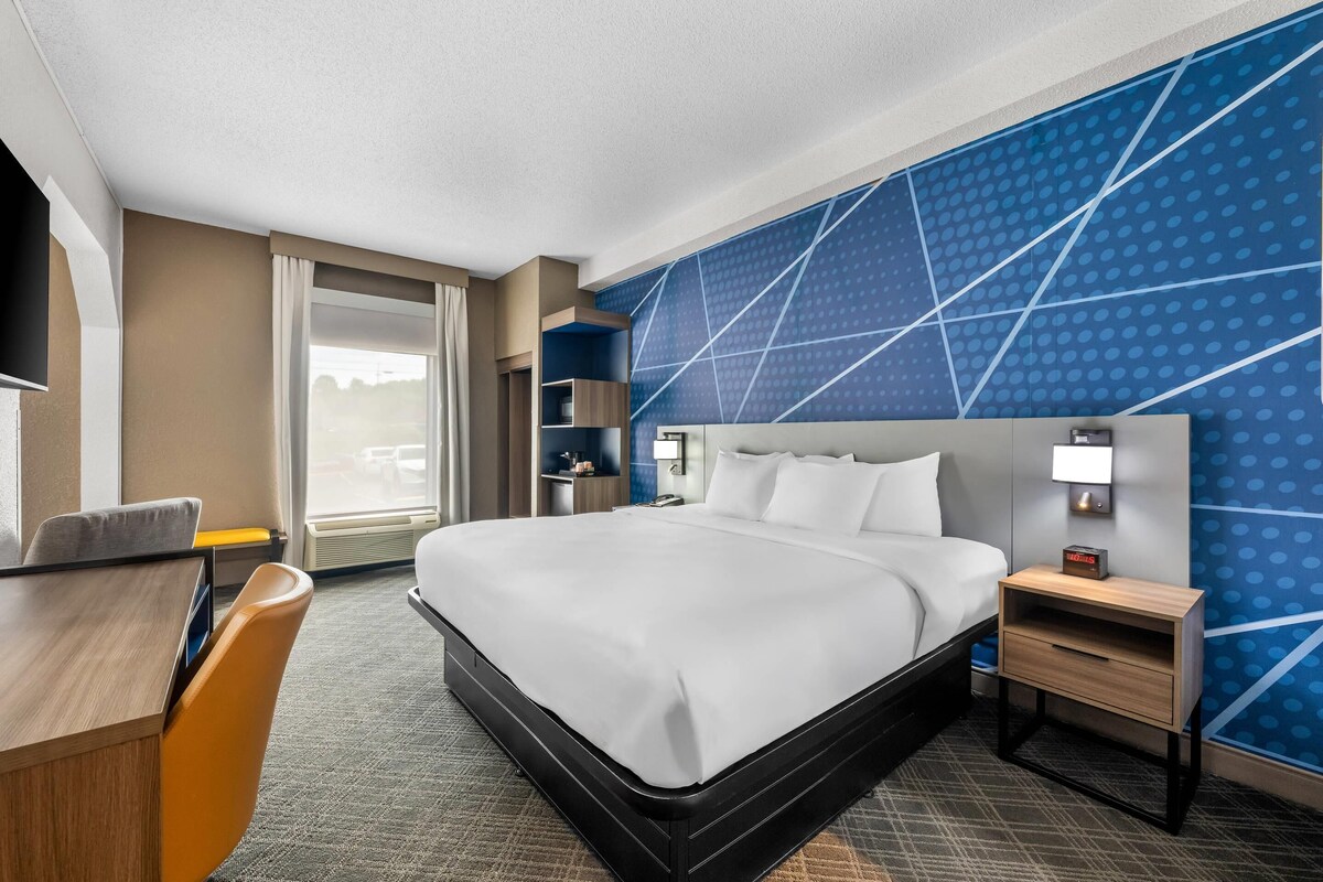 A hotel room with a large bed, a desk, a chair, a nightstand, and a wall with a geometric pattern.