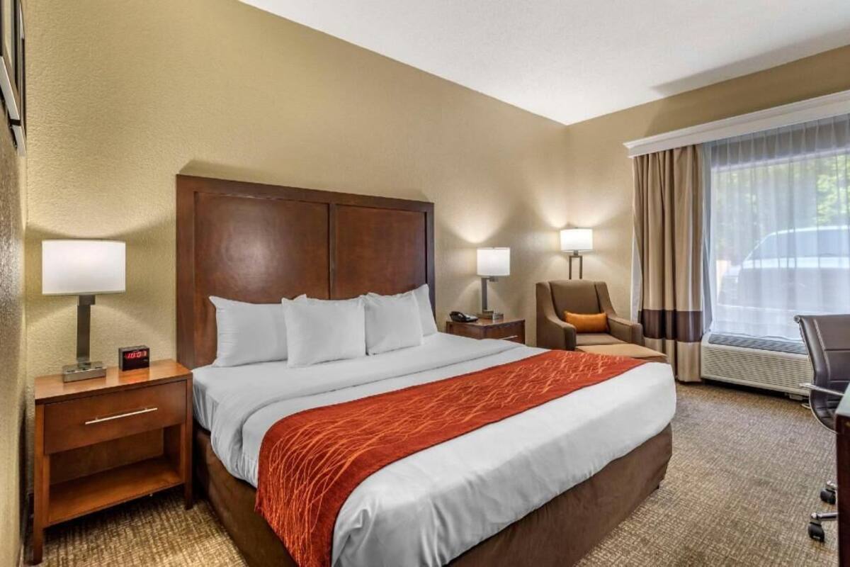 A hotel room with a large bed, two bedside tables with lamps, a chair, a desk, and a large window with curtains.