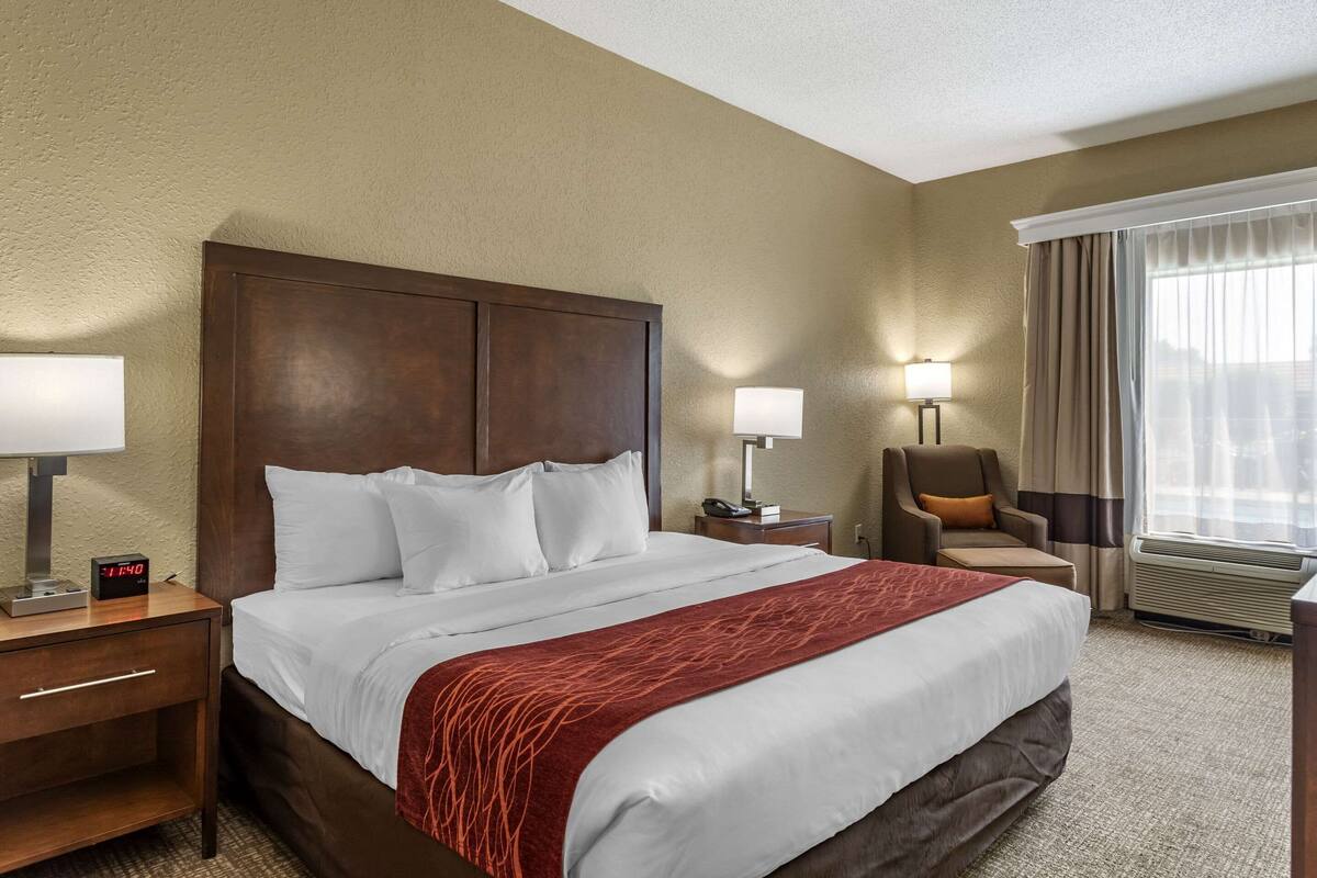 A hotel room with a large bed, a nightstand with a lamp, a chair, and a window with curtains.