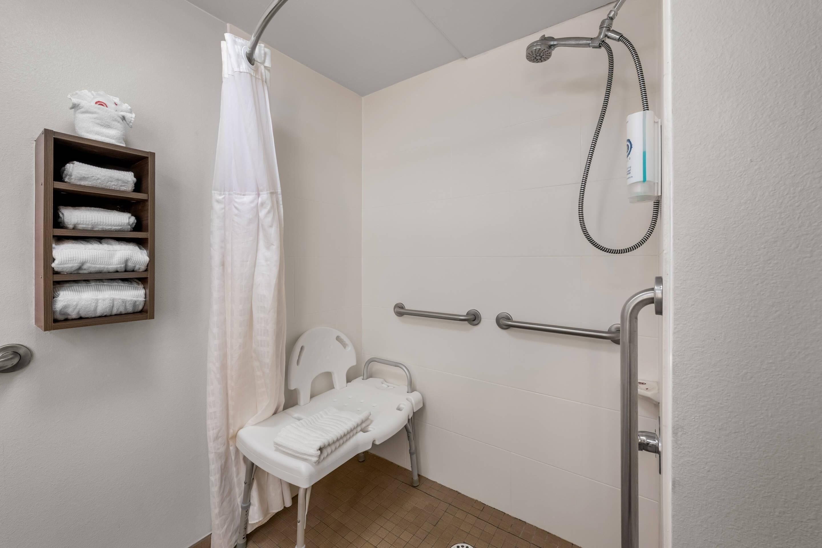 A bathroom with a shower, a white chair, a shelf with towels, and grab bars.
