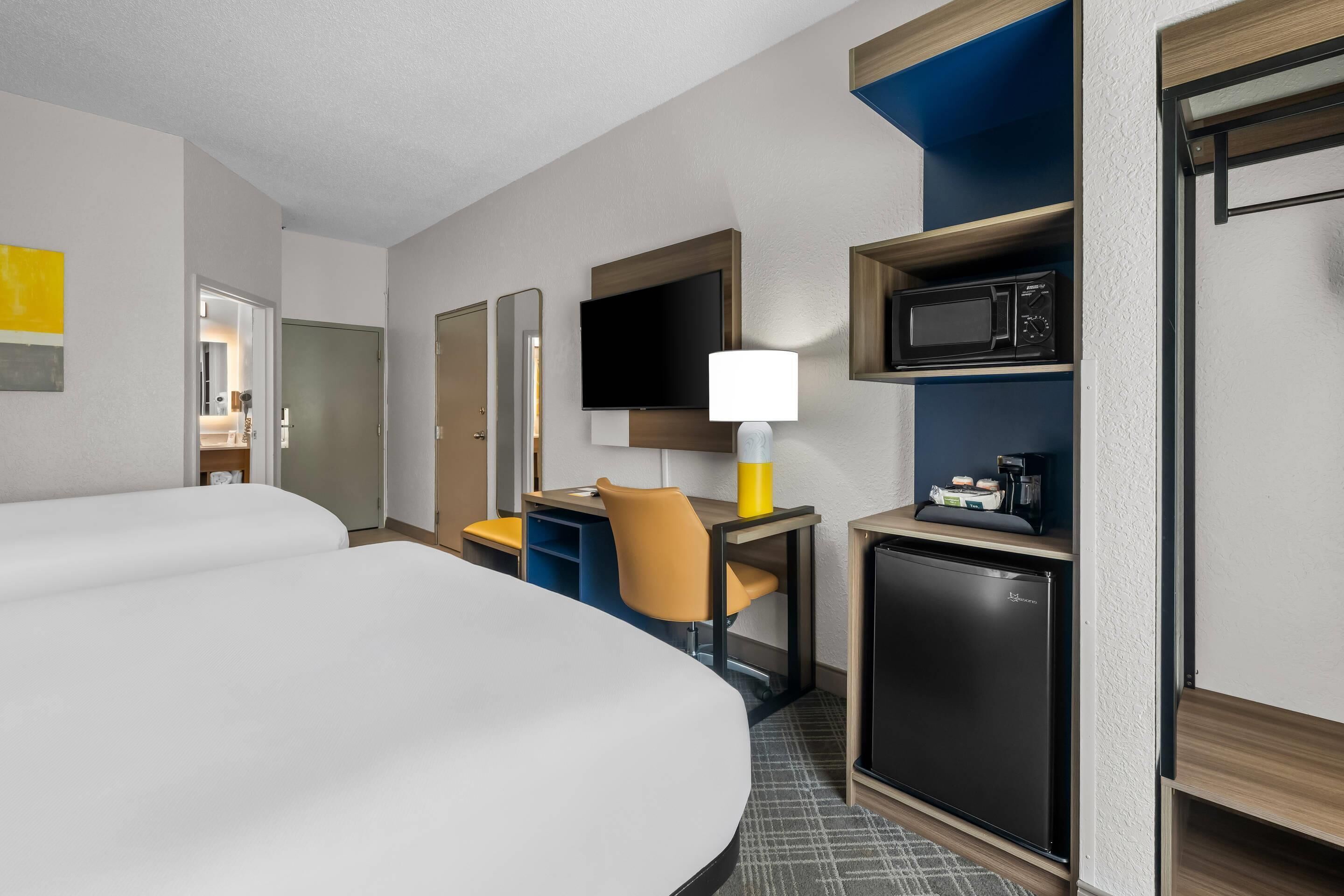 Ein Hotelzimmer mit zwei Betten, einem Schreibtisch mit Lampe, einem Fernseher, einer Mikrowelle und einem kleinen Kühlschrank.