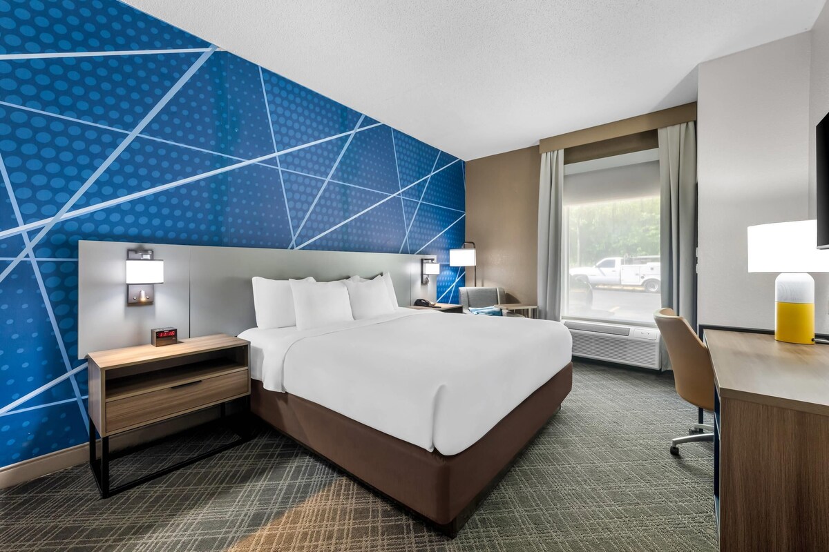 A hotel room with a large bed, a desk, a chair, a nightstand, a lamp, and a view of the outdoors.