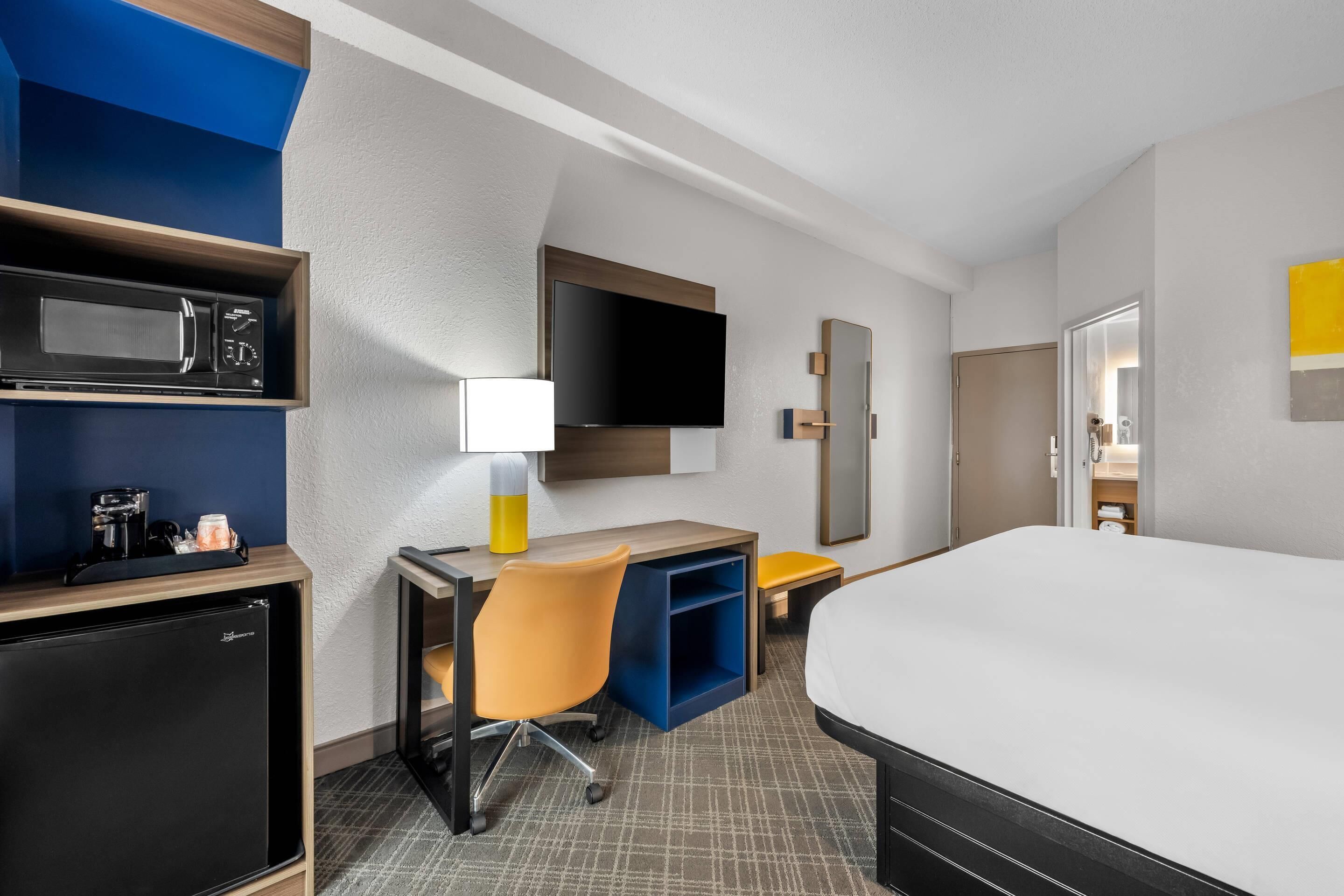 Habitación de hotel con cama, escritorio, silla, microondas, refrigerador, televisor y espejo.