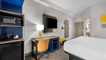Habitación de hotel con cama, escritorio, silla, microondas, refrigerador, televisor y espejo.