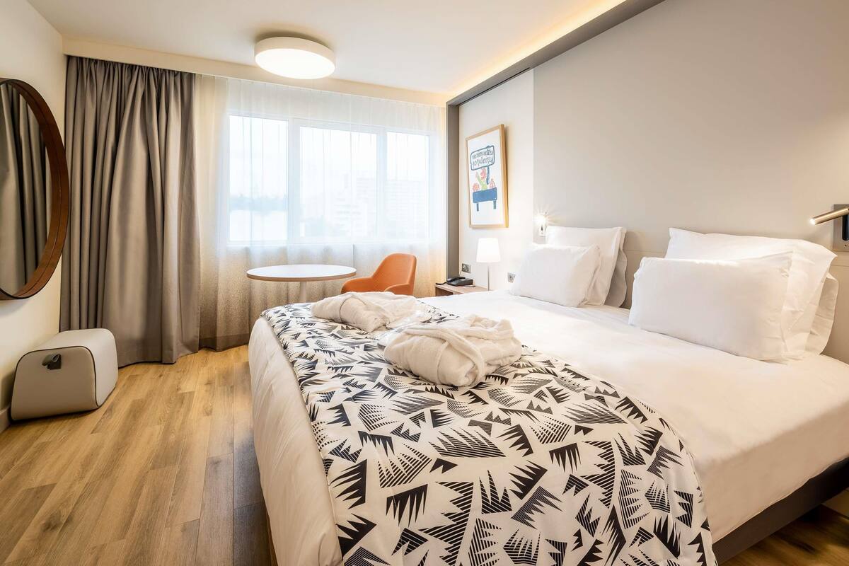 Une chambre d’hôtel comprenant un lit, une table ronde, une chaise, un bureau avec une lampe et un luminaire fixé au mur.