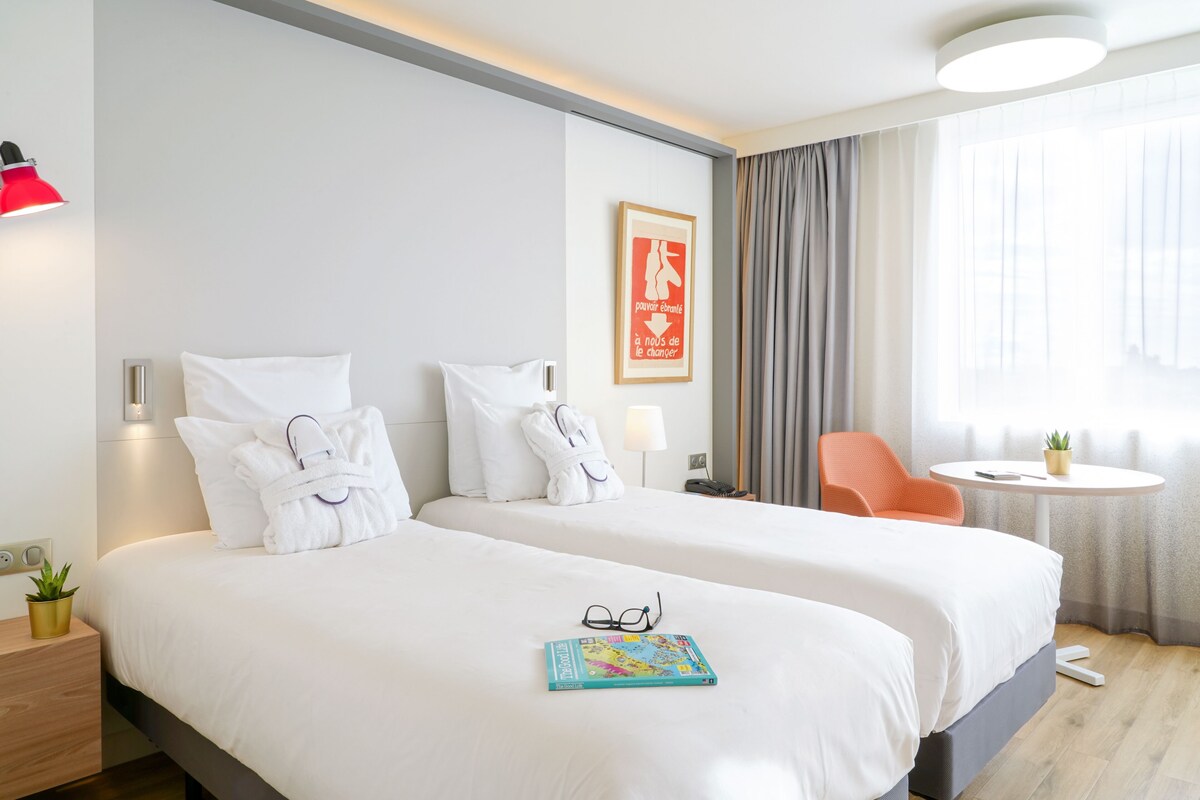 A hotel room with two beds, a small table with a plant, a chair, and a framed picture on the wall.