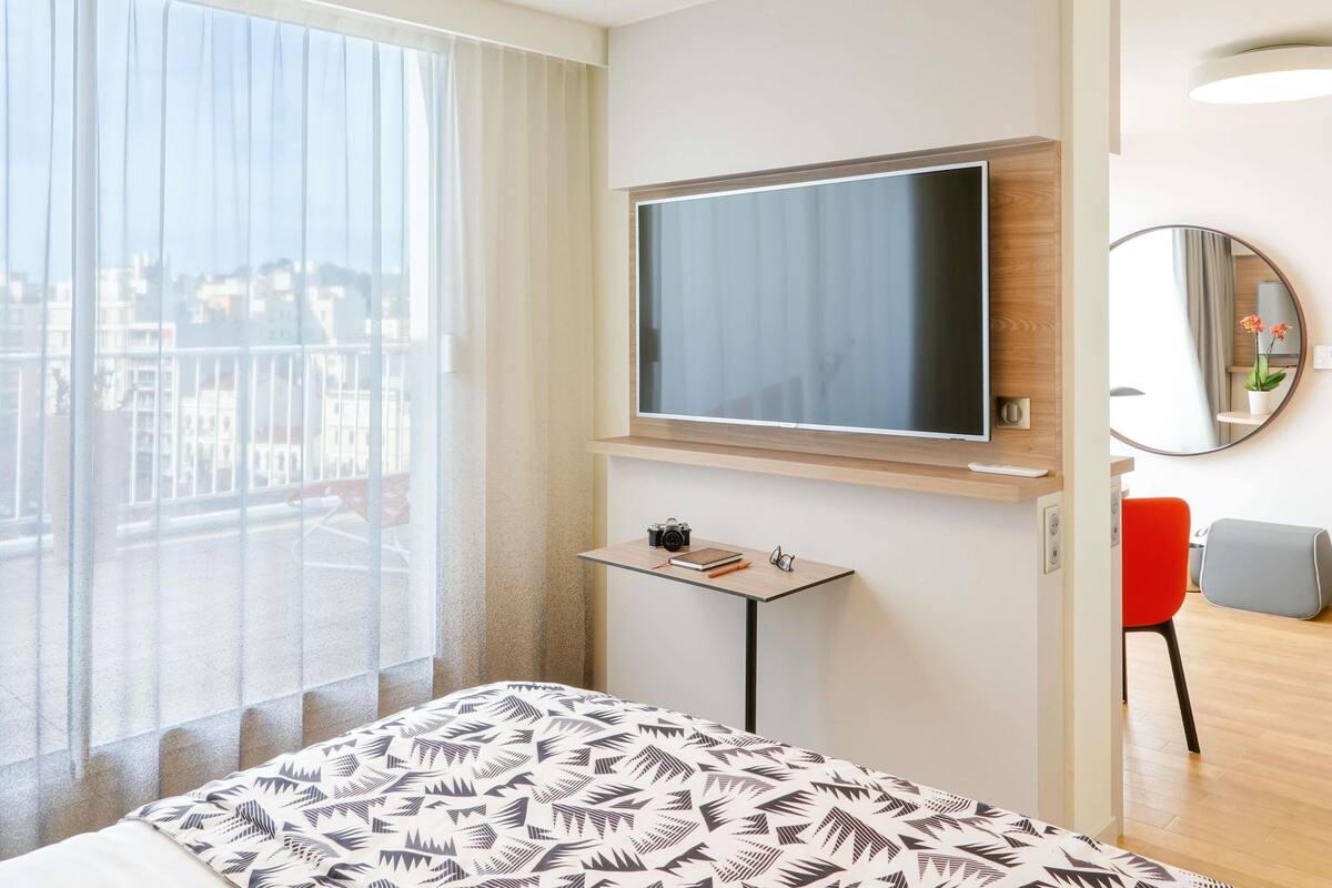 Une chambre d’hôtel avec un grand lit, un téléviseur fixé au mur, une petite table avec un appareil photo, un miroir et une vue sur la ville par la fenêtre.