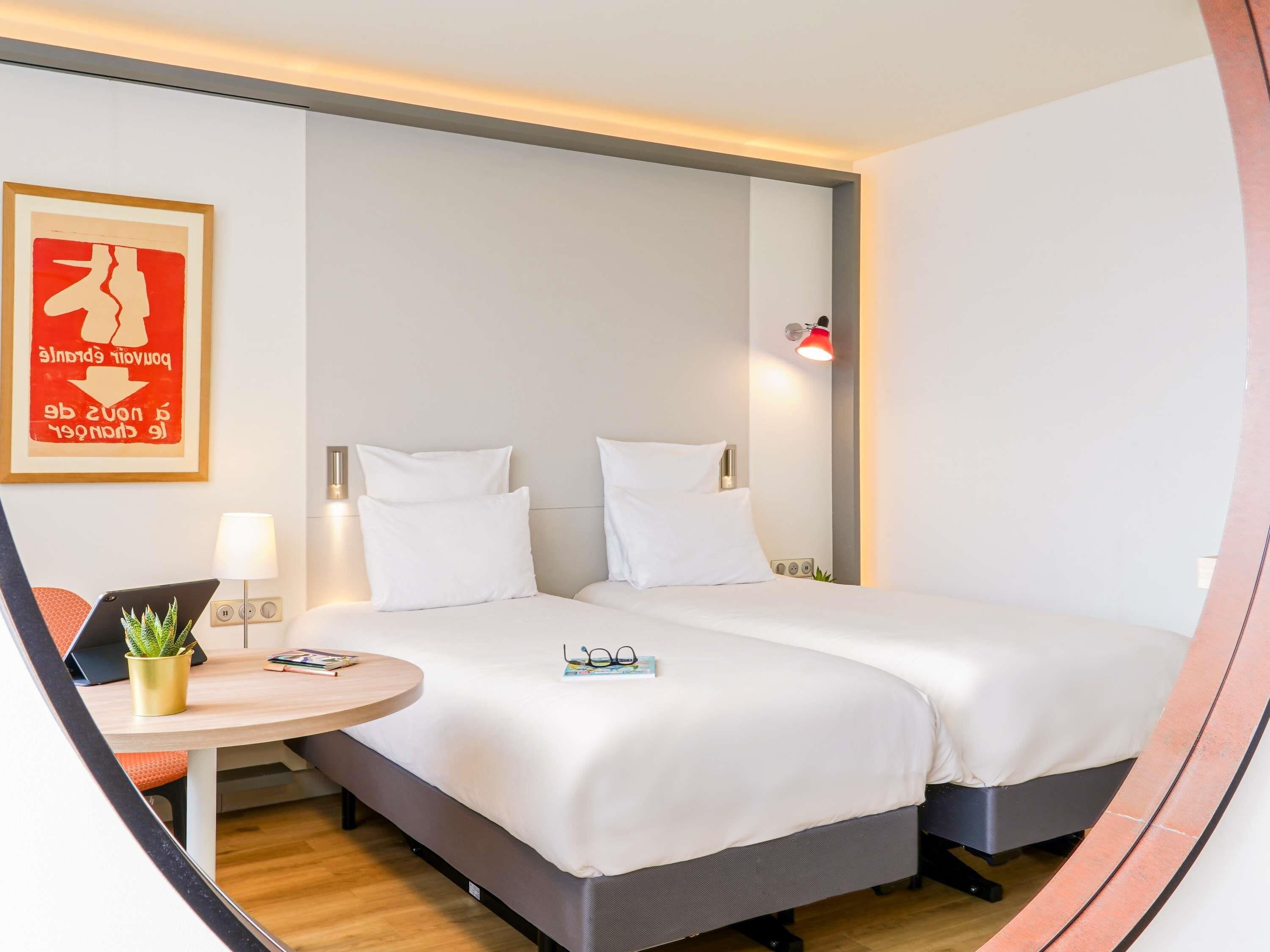Una habitación de hotel moderna con cama, mesitas de noche, un escritorio pequeño y una planta en maceta.