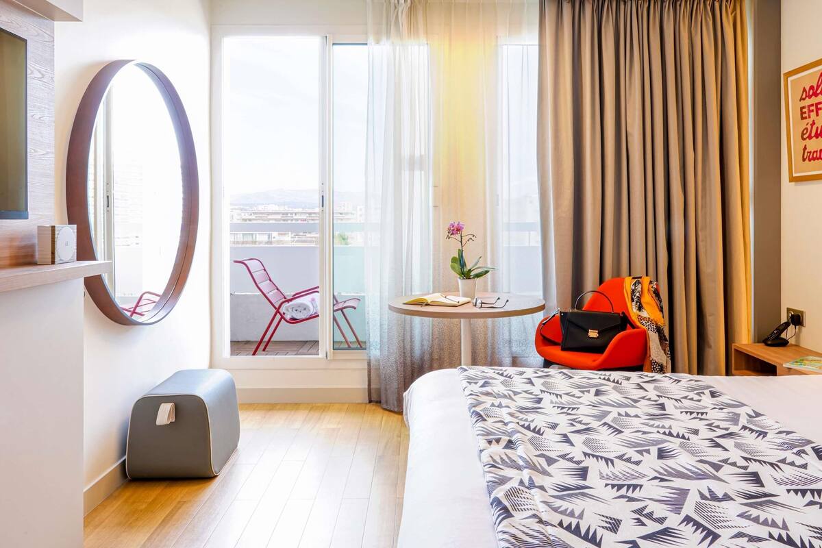 Une chambre d’hôtel avec un lit, un miroir rond, une petite table avec une plante, une chaise rouge et une valise.