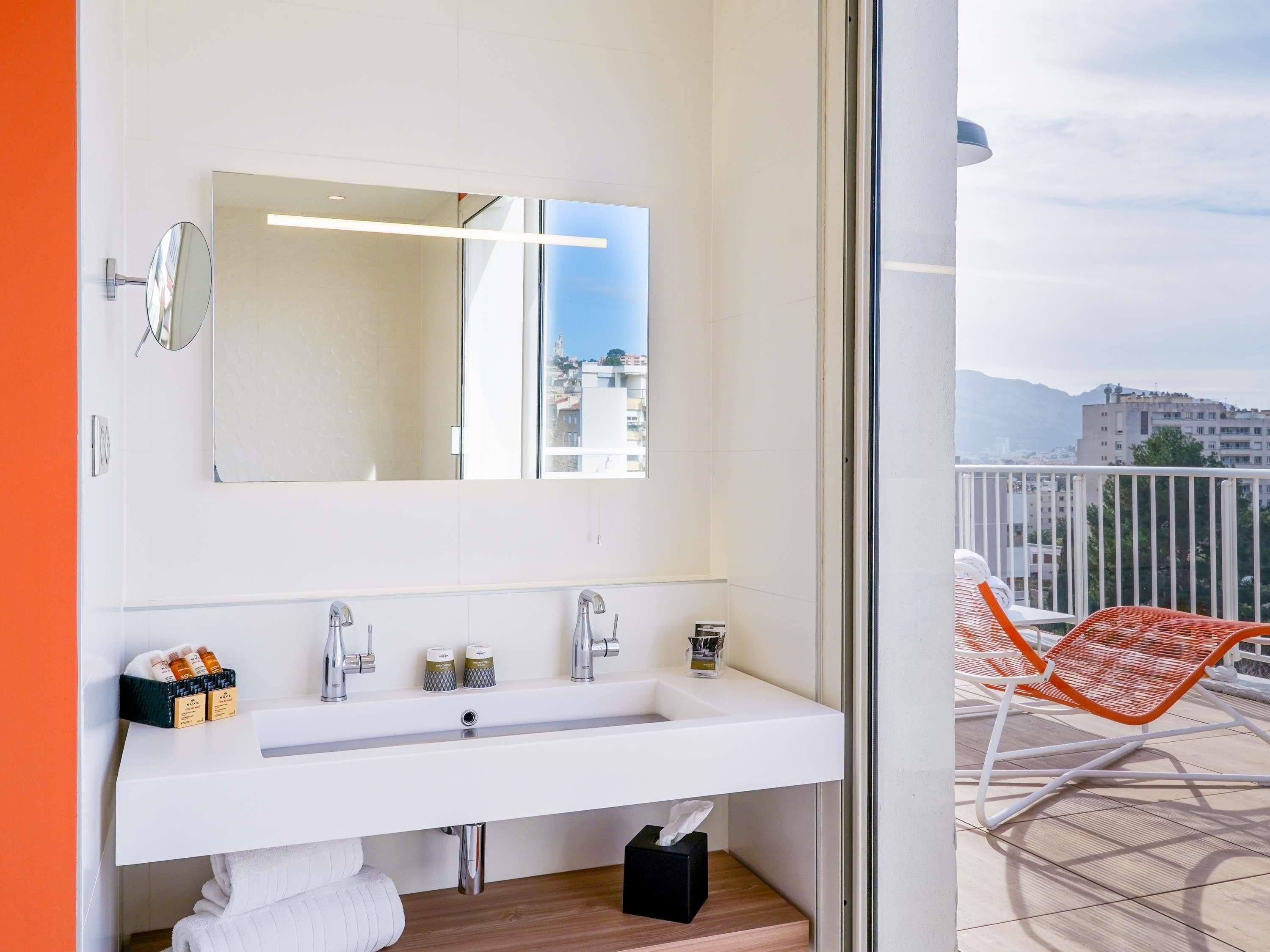 A modern bathroom with a large mirror, a white sink, and a view of the city through a glass door.