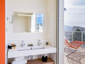 Une salle de bain moderne avec un grand miroir, un lavabo blanc et une vue sur la ville à travers une porte en verre.