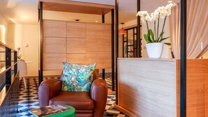 A modern hotel lobby with a brown leather sofa, a colorful floral pillow, a green ottoman, and a wooden reception desk with a potted orchid.