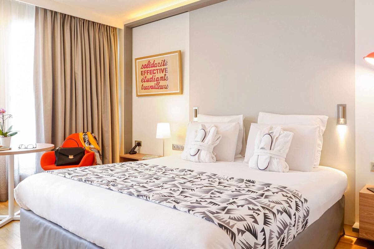 Une chambre d’hôtel avec un grand lit, un bureau avec une chaise, une plante en pot et une citation motivante encadrée au mur.