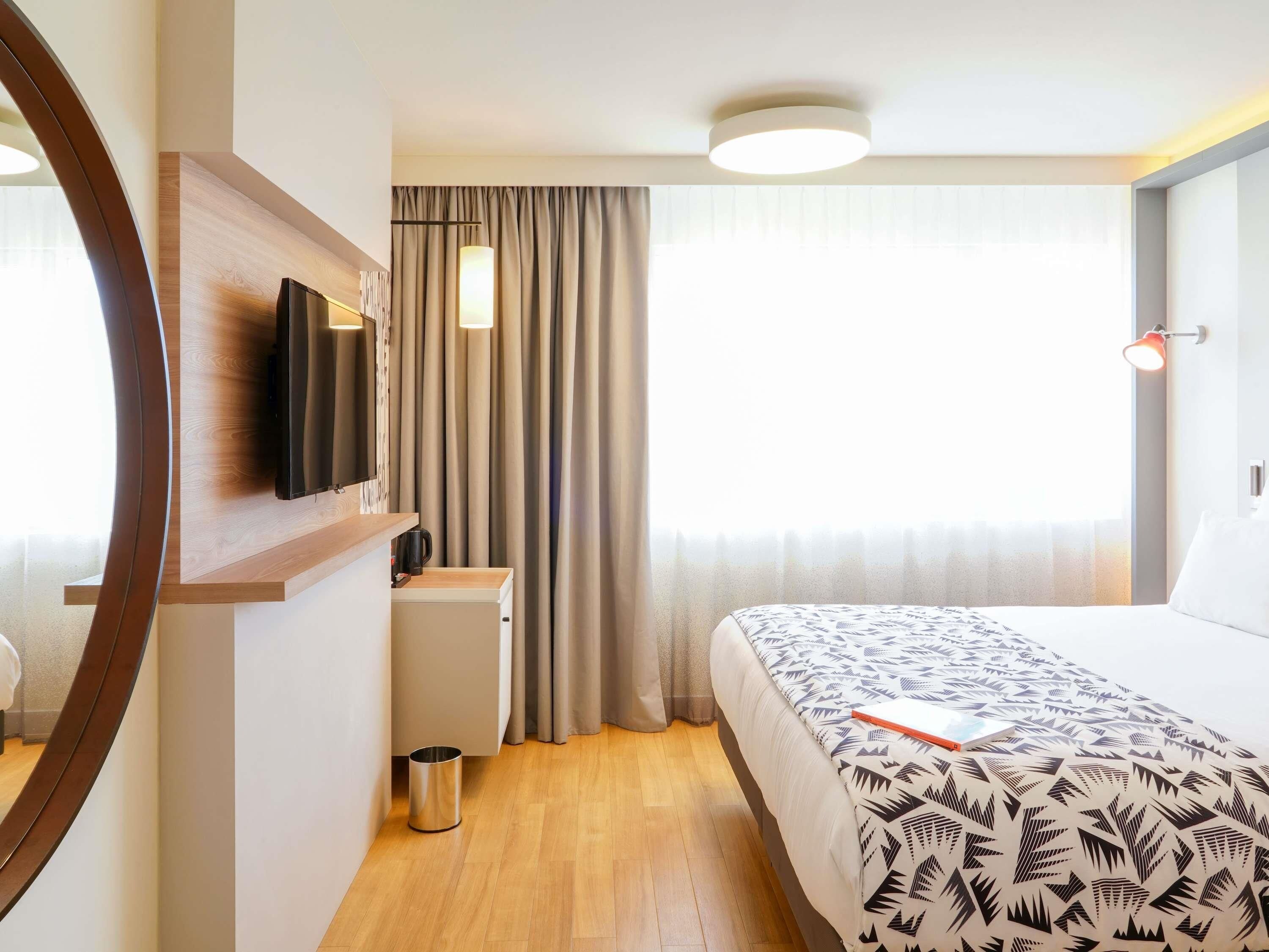 Une chambre d’hôtel avec un lit, un bureau, une télévision, une poubelle et une fenêtre avec des rideaux.