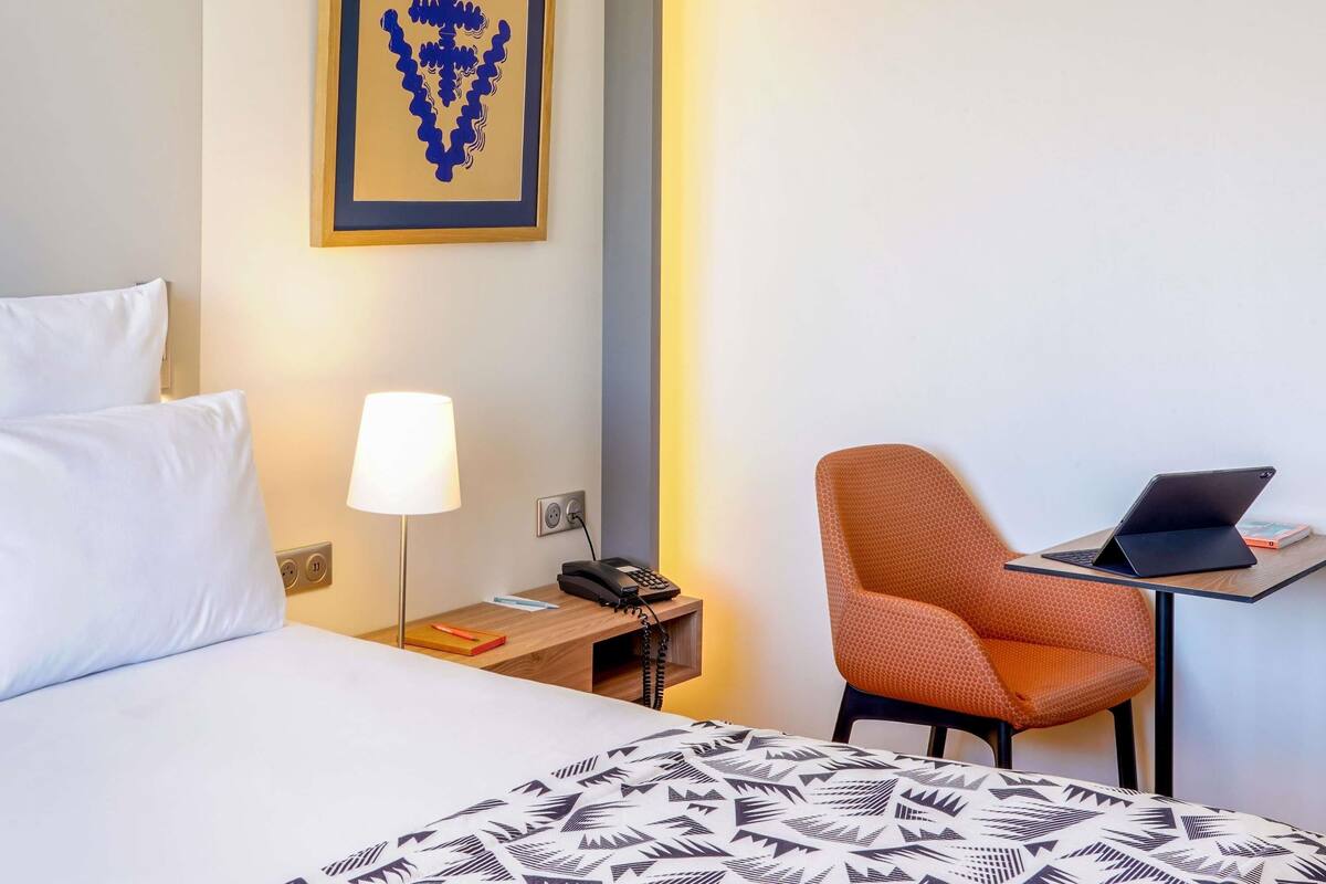 Une chambre d’hôtel équipée d’un lit, d’un bureau avec un ordinateur portable, d’une chaise, d’une lampe et d’un téléphone fixé au mur.