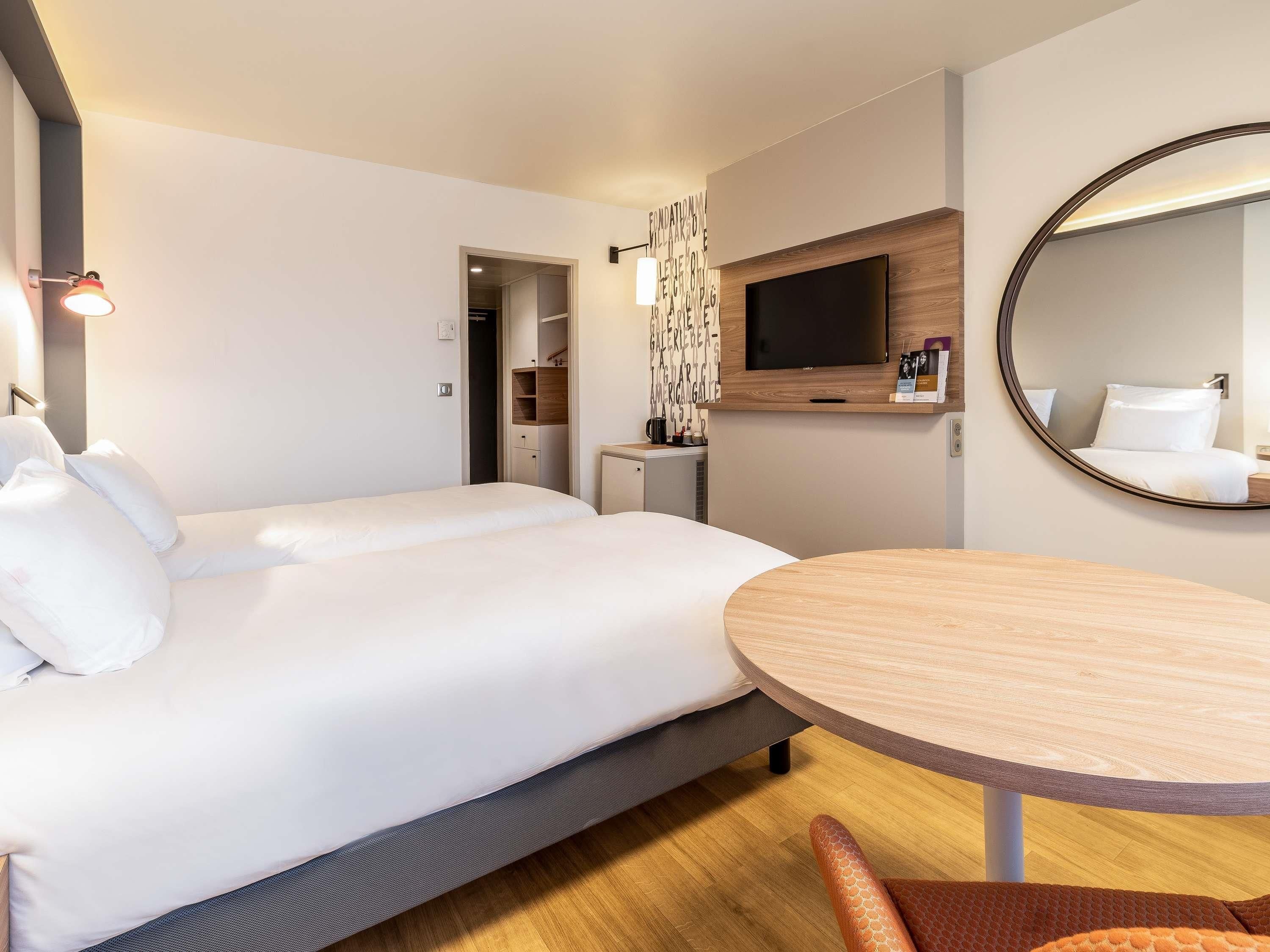 Una habitación de hotel con una cama, una mesa redonda de madera, un televisor y un espejo.
