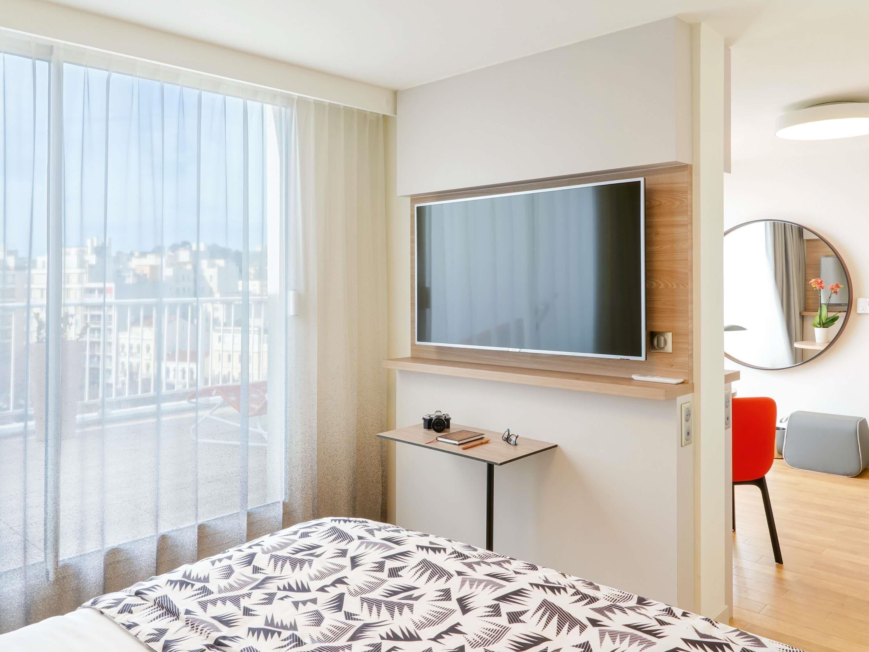 Une chambre d’hôtel avec un grand lit, un téléviseur fixé au mur, une petite table avec un appareil photo et des livres, un fauteuil rouge et une vue sur la ville grâce à de grandes fenêtres.