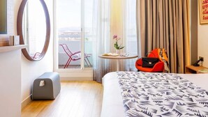 Hotellihuone, jossa on sänky, työpöytä, tuoli, pieni pöytä, jossa on kasvi, matkalaukku ja ikkunasta avautuva näkymä kaupunkiin.