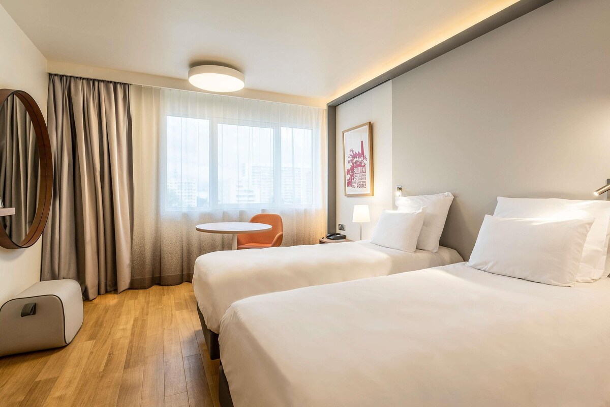 A hotel room with two beds, a round table, a chair, a lamp, and a framed picture on the wall.