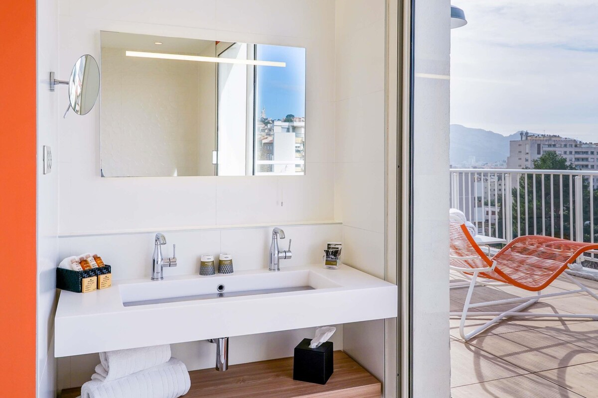 Un baño moderno con un espejo grande, un lavabo blanco y vistas de la ciudad a través de una puerta de cristal.