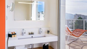 A modern bathroom with a large mirror, a white sink, and a view of the city through a glass door.