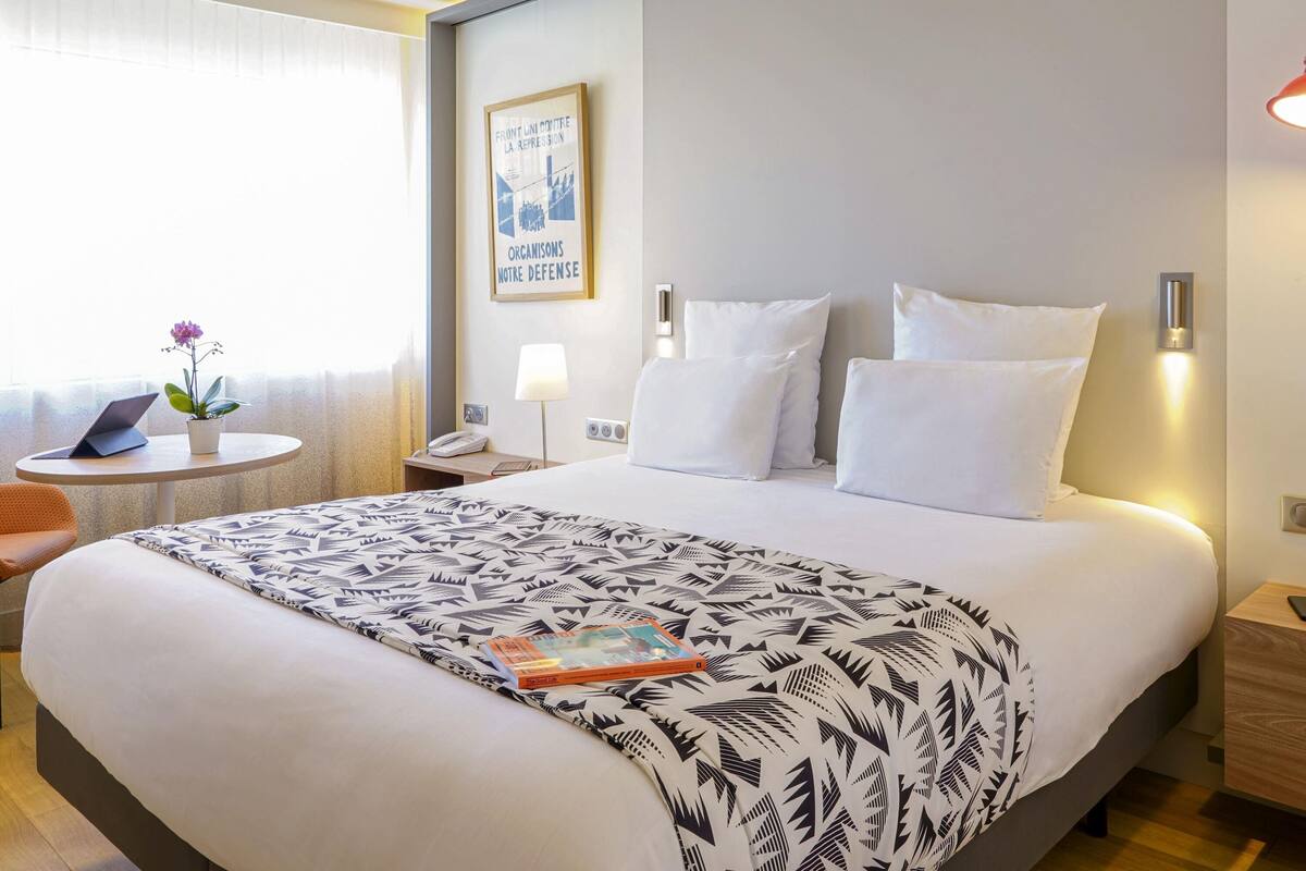 Une chambre d’hôtel avec un lit, des tables de chevet, une lampe, une petite table avec un ordinateur portable et une plante, et une affiche accrochée au mur.