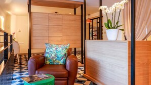 Un moderno vestíbulo de hotel con un sofá de cuero marrón, un cojín floral colorido, un reposapiés verde y un mostrador de recepción de madera con una orquídea en maceta.