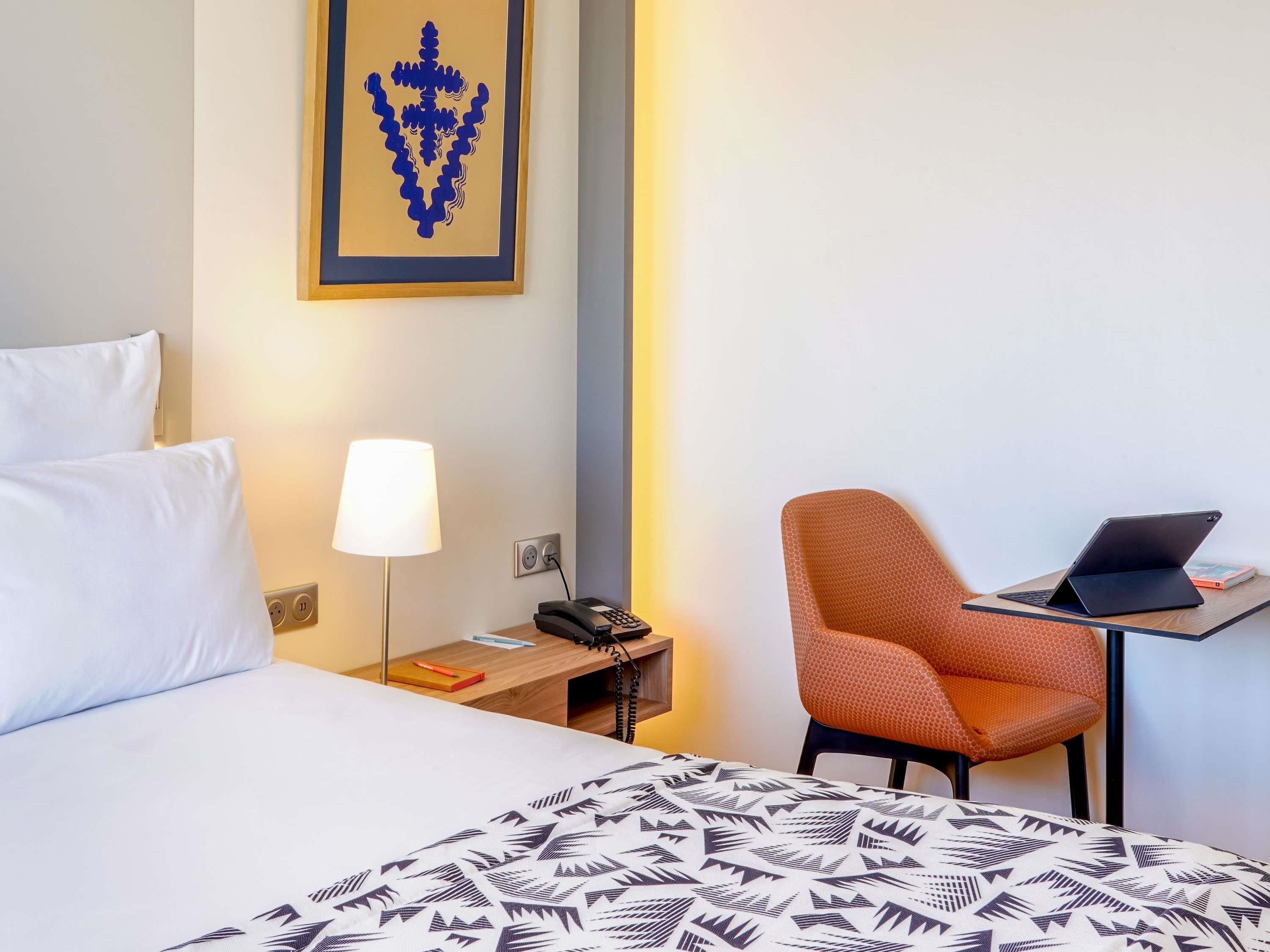 A hotel room with a bed, a desk with a laptop, a chair, a lamp, and a wall-mounted phone.