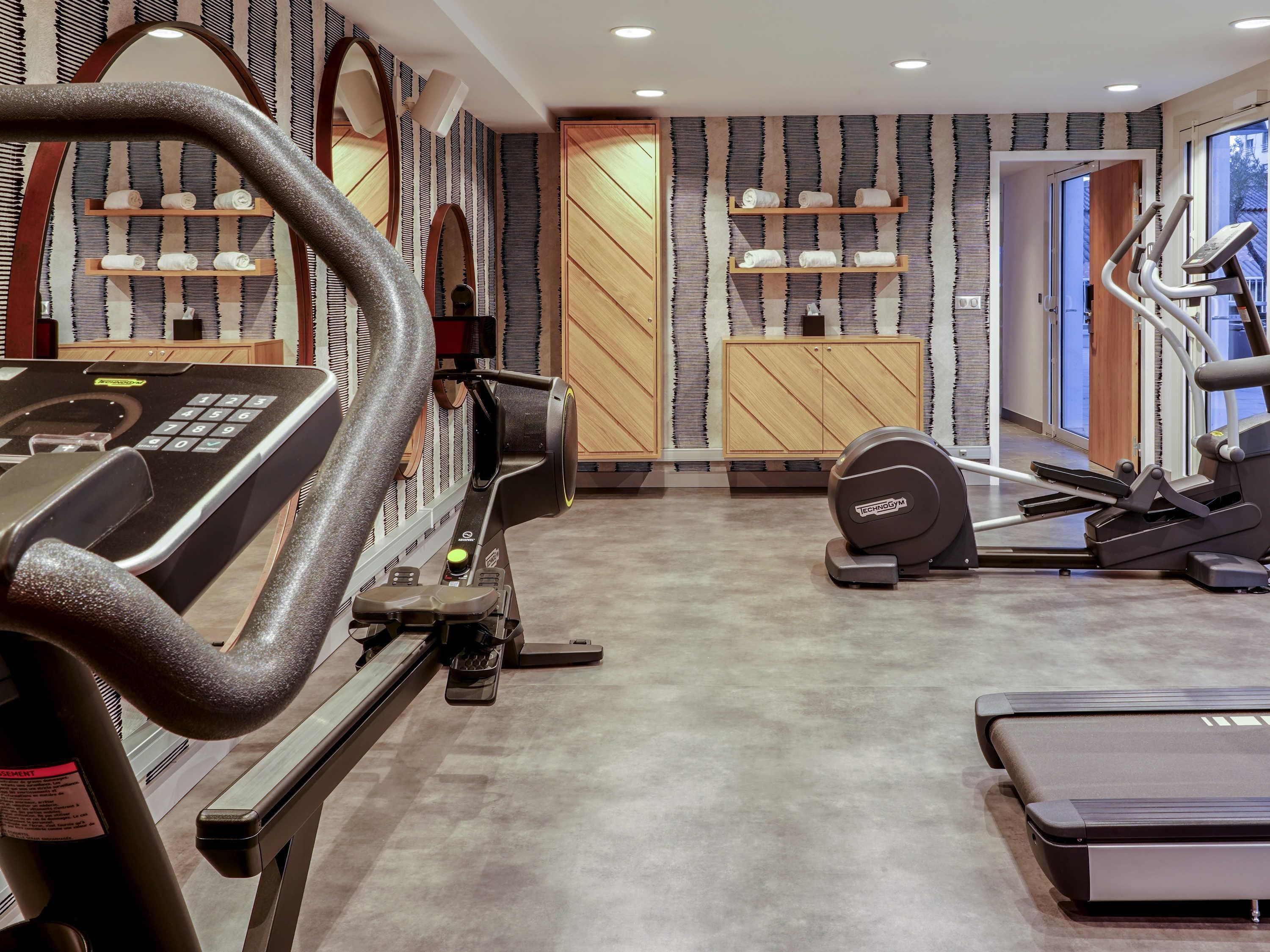 Une salle de sport équipée de matériel d’exercice, avec des étagères en bois où sont rangées des serviettes et un mur décoré de motifs.
