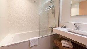 Moderni kylpyhuone, jossa on lasiseinällä varustettu suihku, valkoinen kylpyamme ja puinen allaskaappi, jossa on peili.