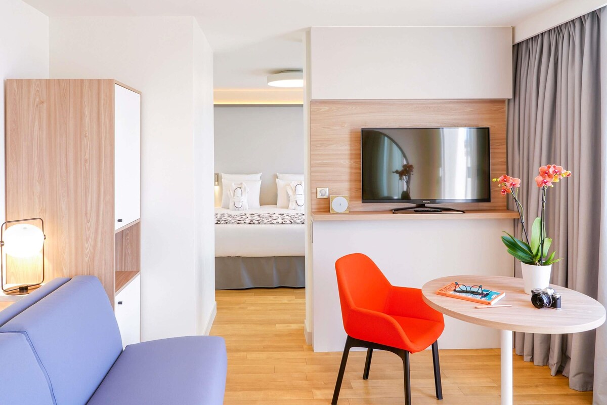 A modern hotel room with a sofa, a round table with a chair, a TV, and a bed with a view.